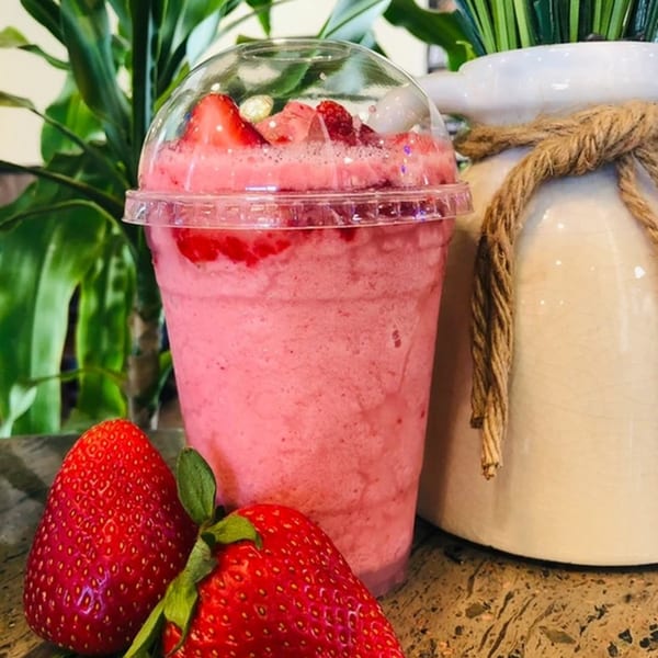 A vibrant pink strawberry smoothie in a clear cup, garnished with fresh strawberries and lychee, beside whole strawberries.