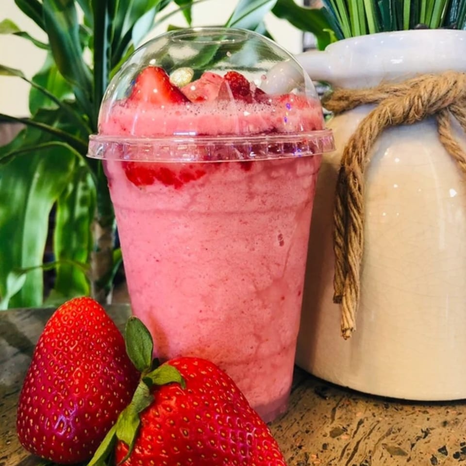 A vibrant pink strawberry smoothie in a clear cup, garnished with fresh strawberries and lychee, beside whole strawberries.