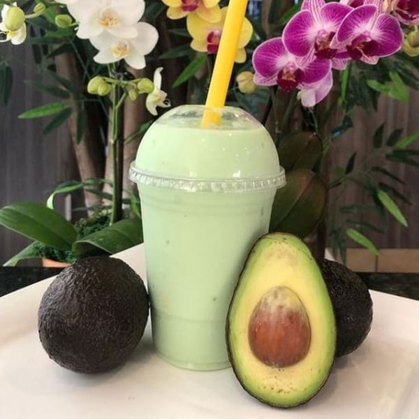 Avocado smoothie with a yellow straw, whole avocados, and a halved avocado on a white plate.